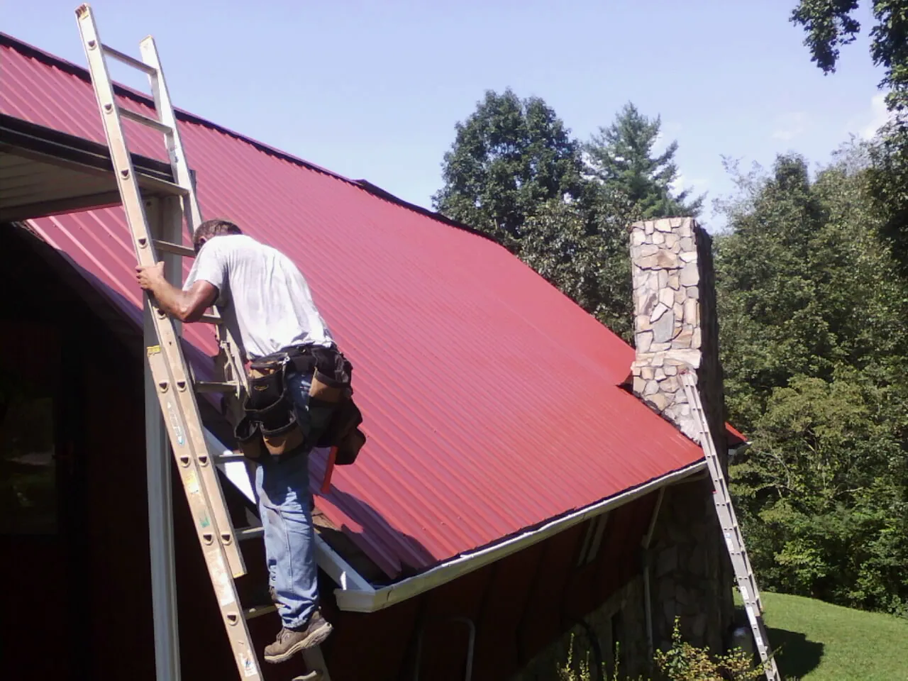 Roofer on a ladder beside a red metal roof and stone chimney — Soffit & Fascia Repair by Roofing Brothers in Delavan